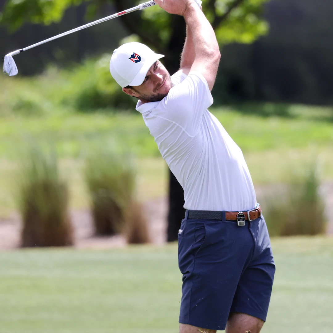 Man in white shirt and cap swinging a golf club on a green field, with trees in the background.