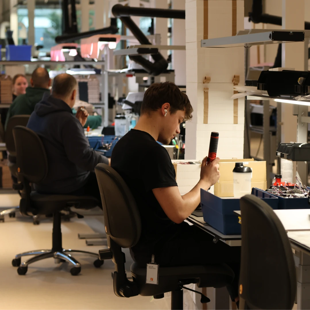 People working at desks in a busy workshop, assembling equipment with tools and materials around them.