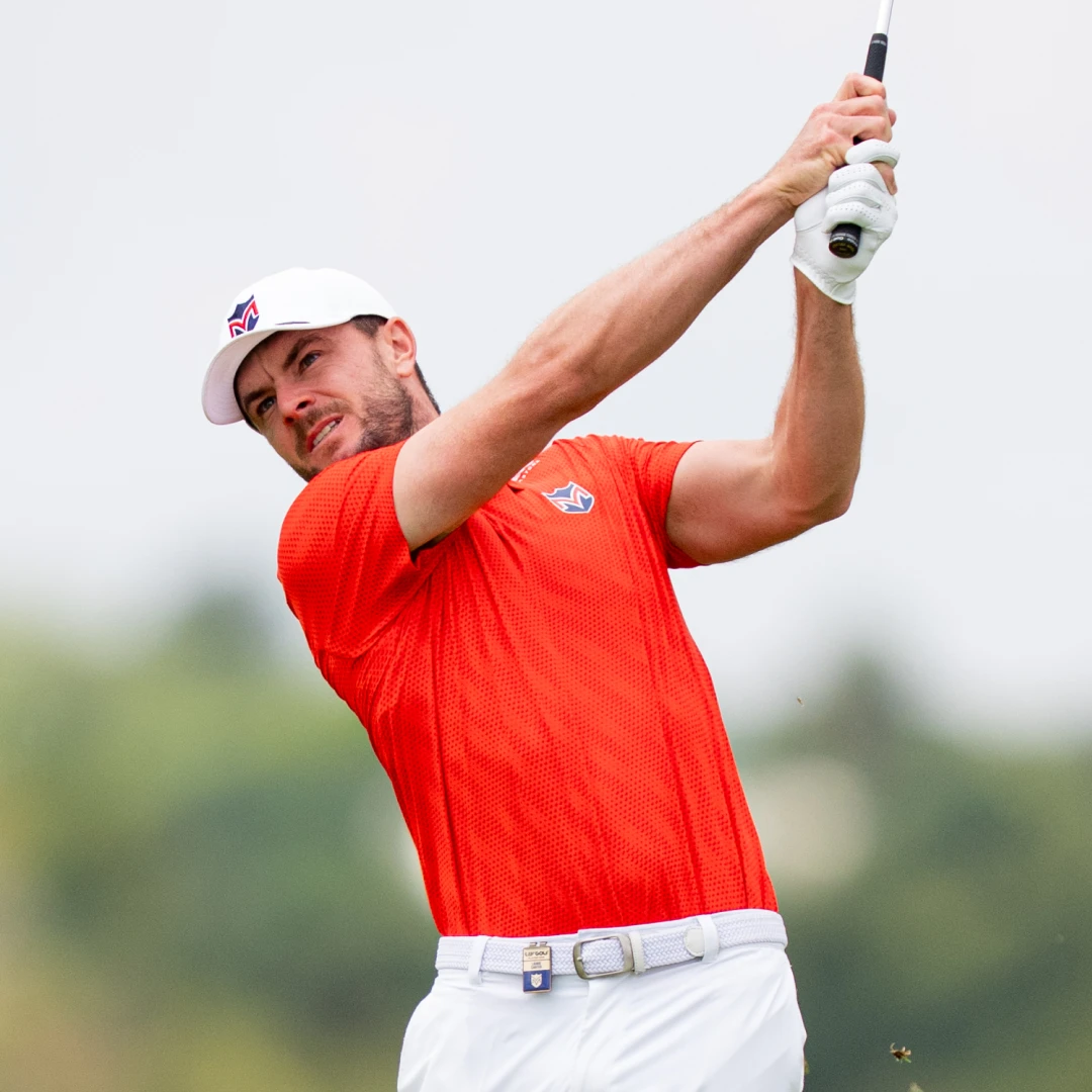 Golfer in a red shirt and white cap swinging a club against a blurred green background.