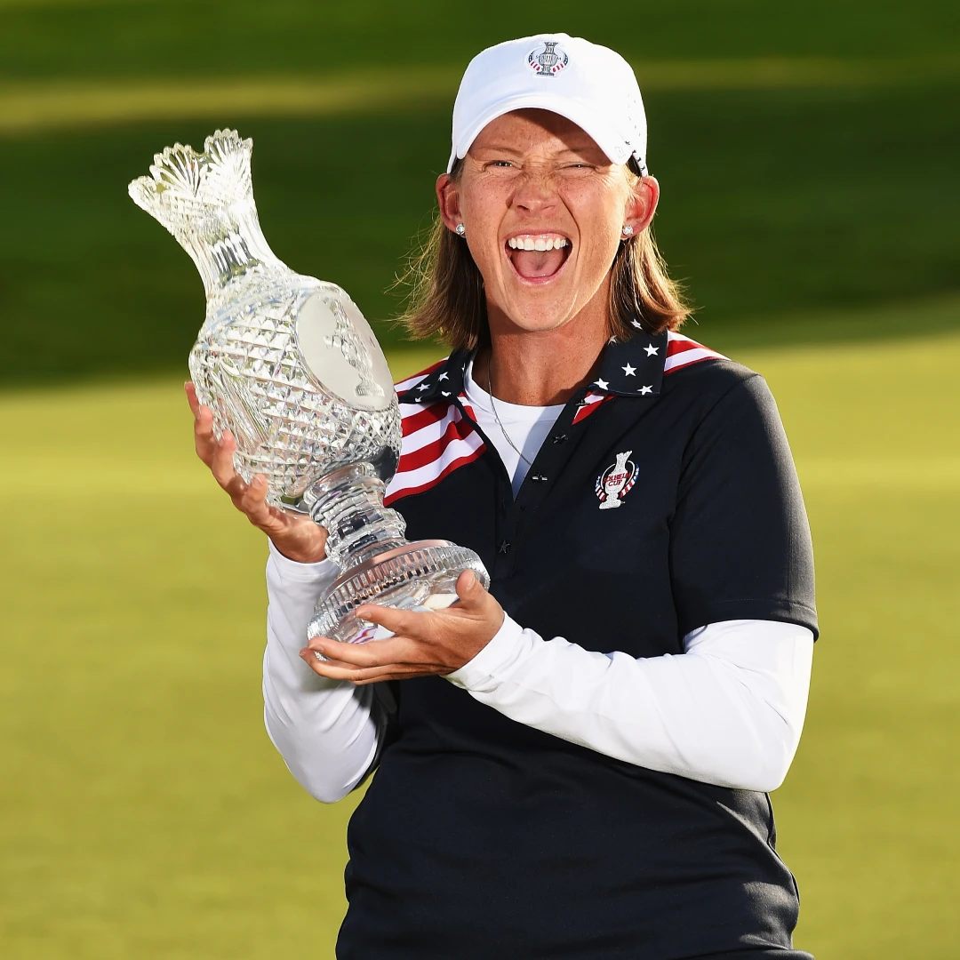A person in a navy and white outfit holding a crystal trophy, smiling widely, with a grassy background.
