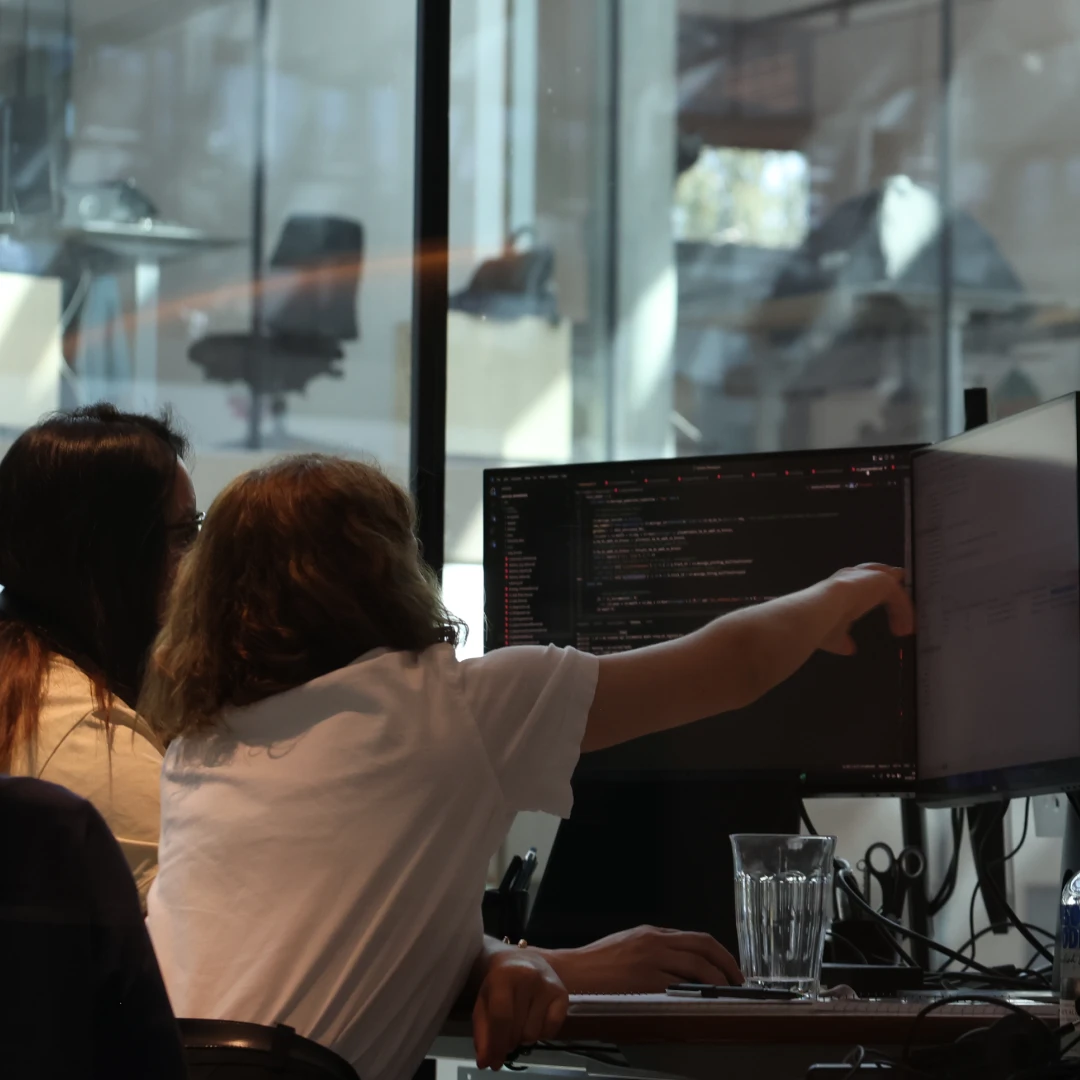 Two people collaborate in an office, focusing on code displayed on a dual-monitor setup. One points at the screen, discussing the work.