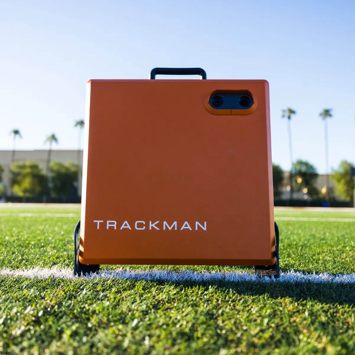 Orange TrackMan device with dual lenses and handle resting on a turf field white line, blurred palm trees and buildings in background.