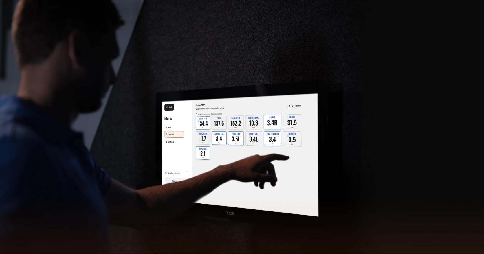 A person points at a touchscreen displaying golf data metrics in a dimly lit room.