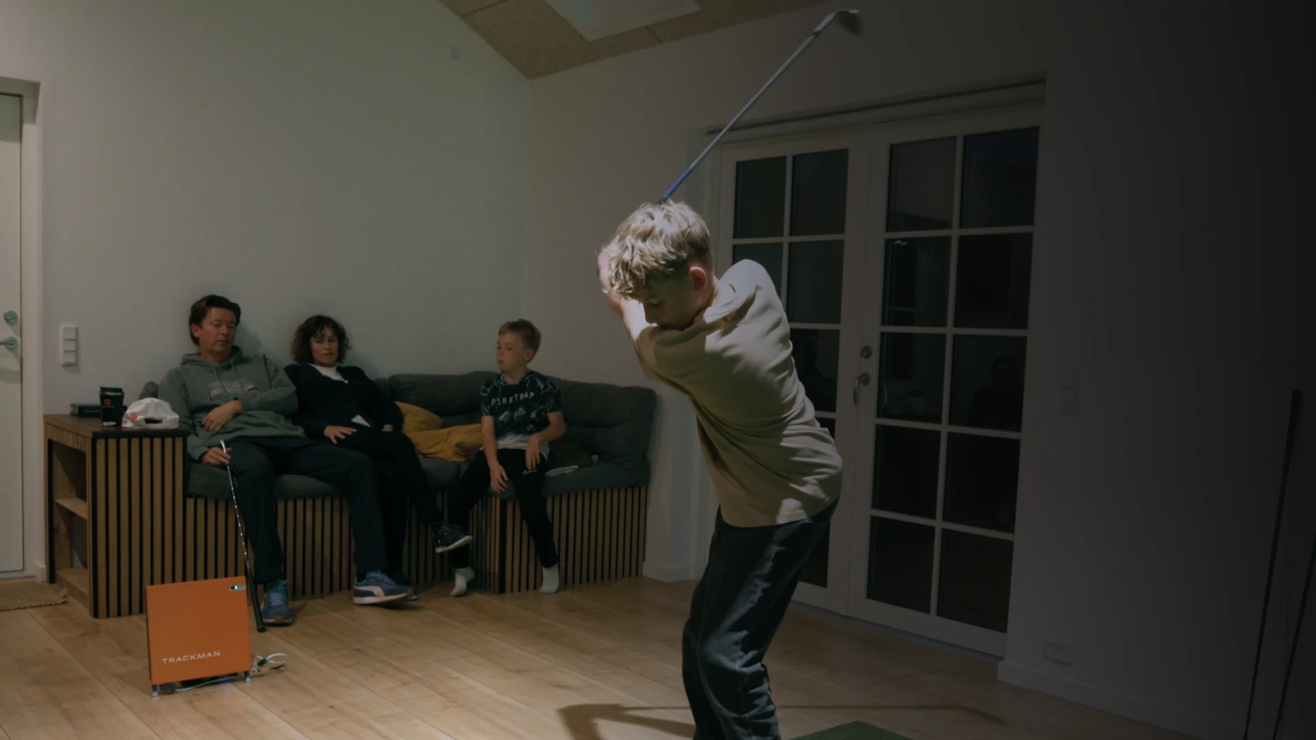 Boy practicing golf swing indoors while two others sit on a couch watching. Room is dimly lit with wooden floor and large window.
