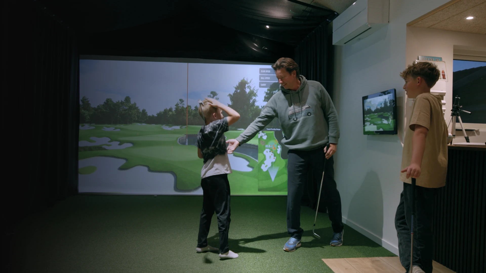 A man and a child play indoor golf, with a screen projecting a golf course. The child uses a golf club while the man watches and gestures.
