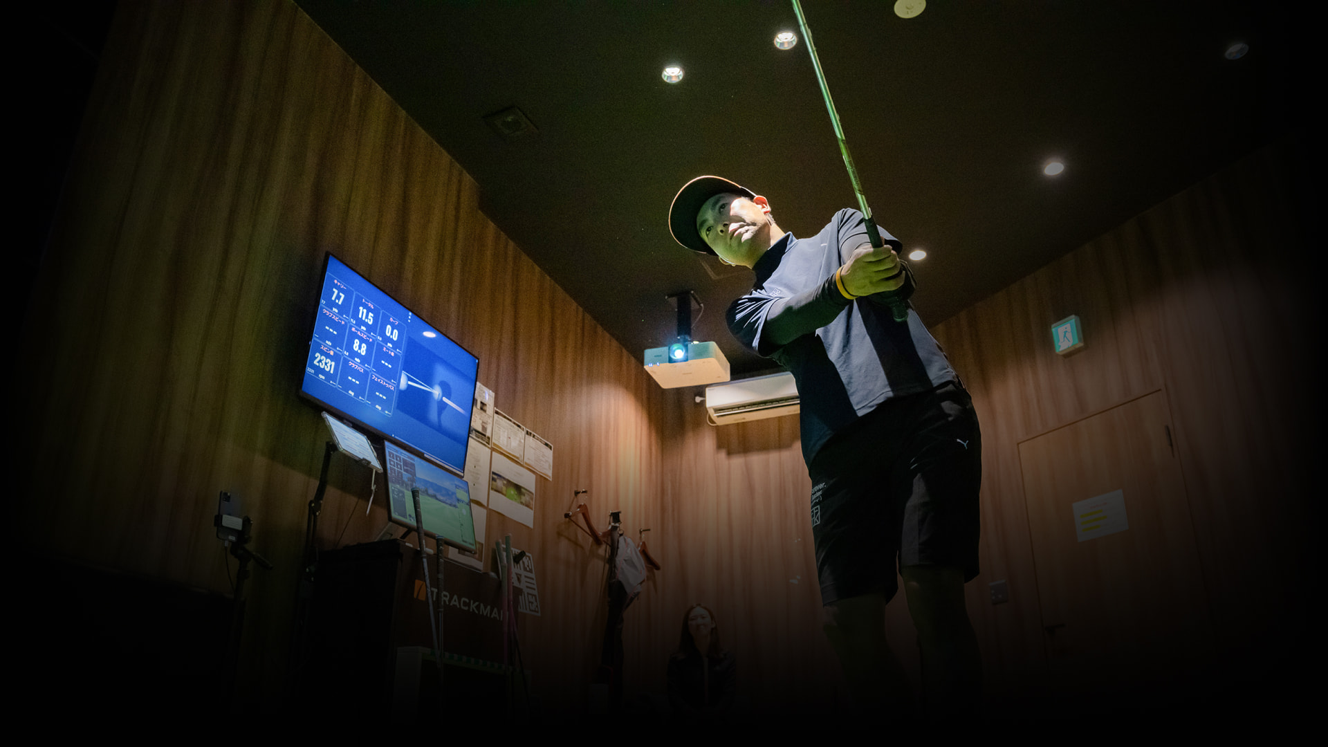 Person practicing golf swing indoors, wearing a cap and dark clothing, with a screen displaying data in the background.