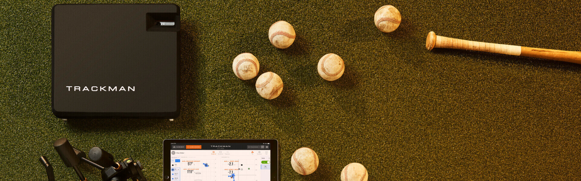 Trackman Baseball equipment on grass: Trackman device, tablet displaying data, camera, wooden bat, glove, and several baseballs.