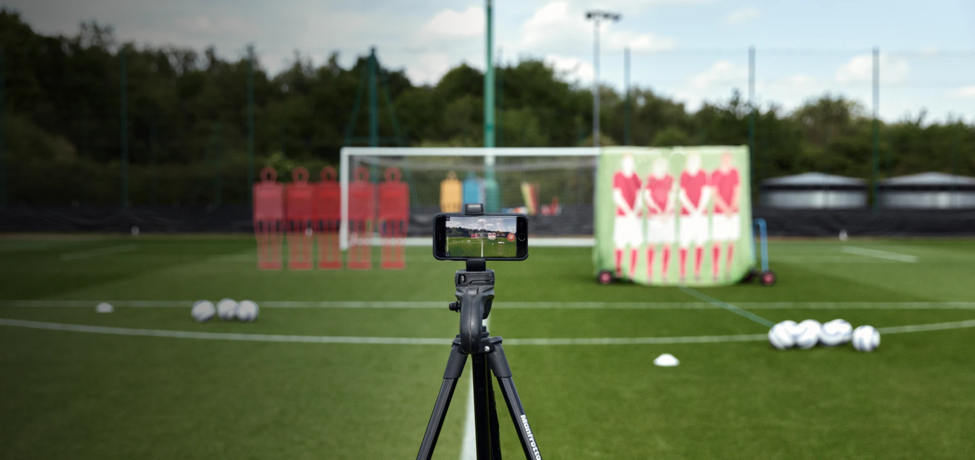 A smartphone on a tripod records a soccer training session with mannequins and balls on a field.