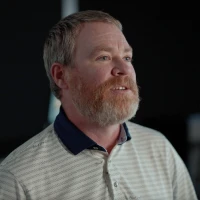 A bearded man in a striped polo shirt looks to the side against a dark background.