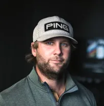 A person wearing a gray cap with "PING" on it and a green zip-up jacket stands against a dark background, looking towards the camera.