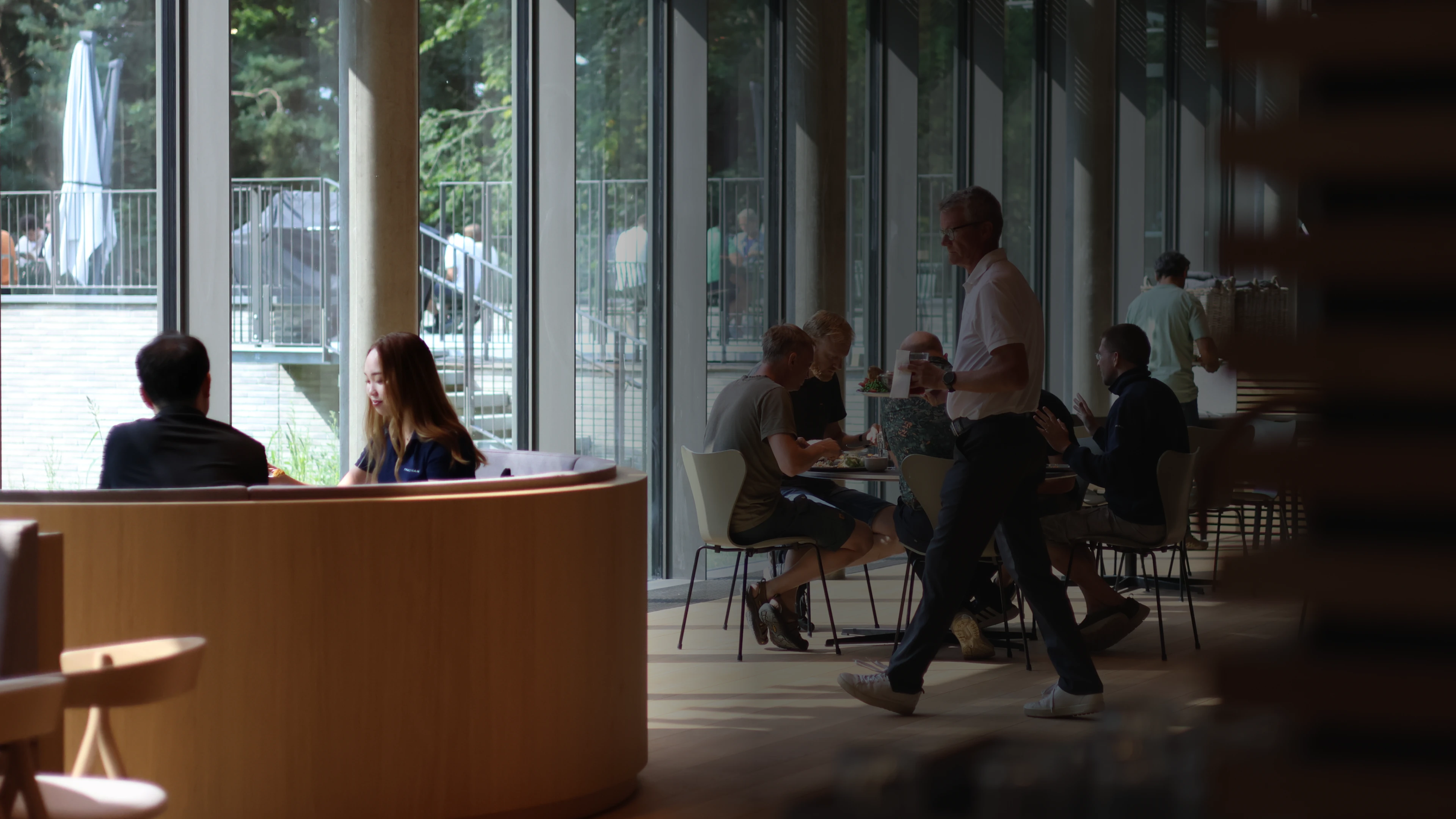 People sitting and conversing in a sunlit cafe with large windows and trees visible outside.