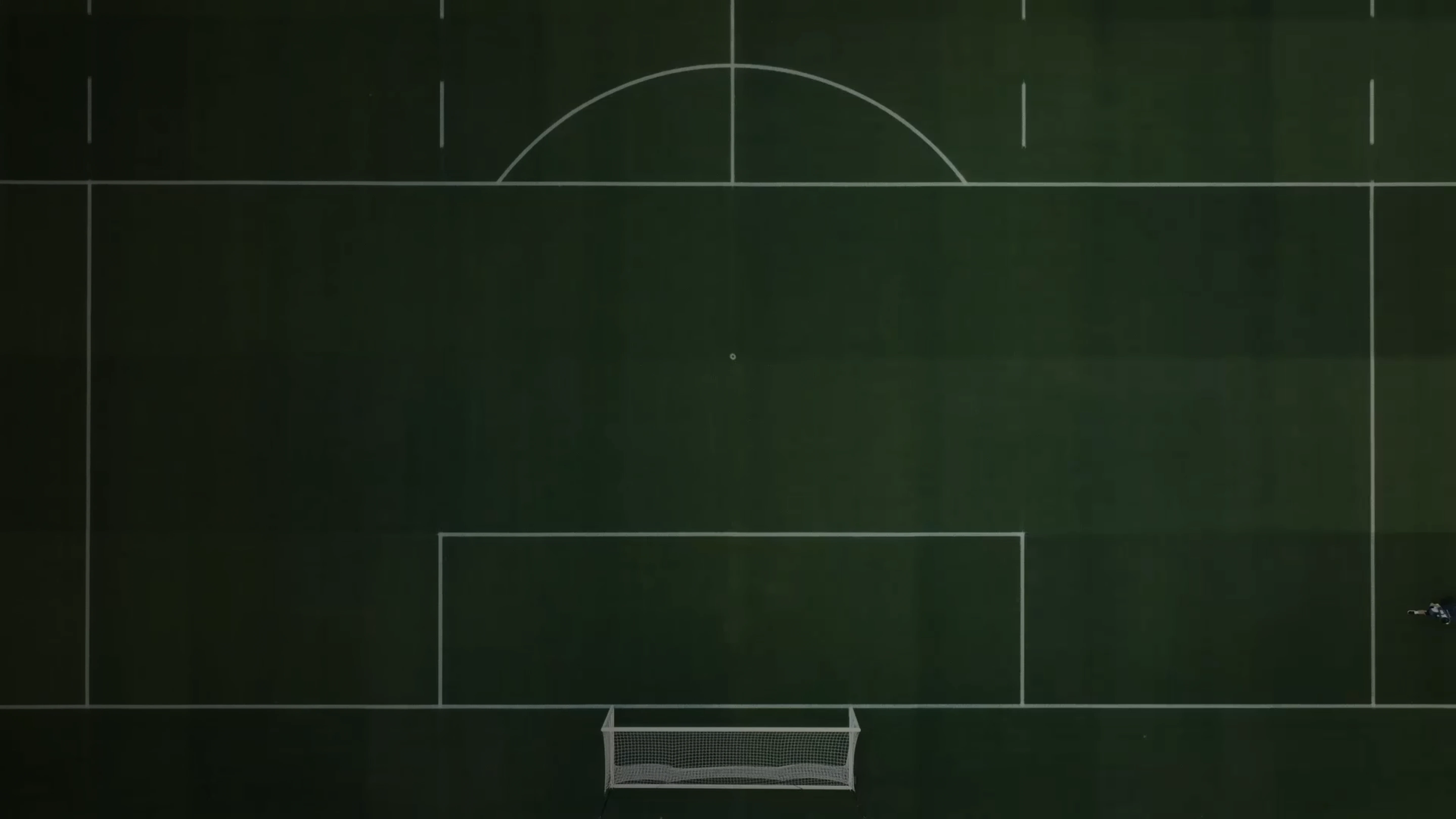 Aerial view of a dark green soccer field with white lines, a centered goalpost, and a single player in motion near the bottom right corner.
