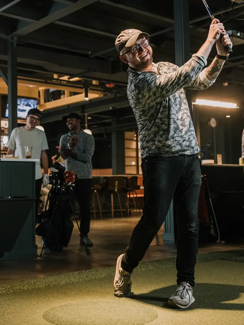 A man in a golf pose indoors, smiling, with onlookers and golf equipment in the background. Indoor setting with modern decor.