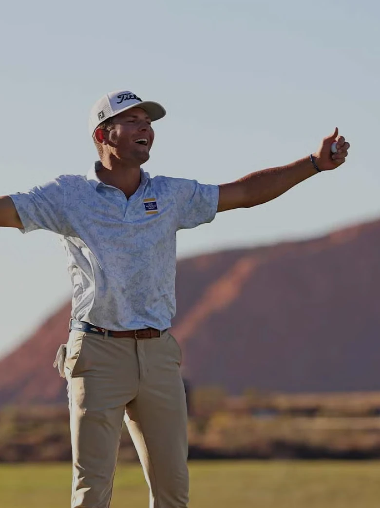Golfer in a light shirt and beige pants joyfully raises arms on a course with scenic hills and blue sky in the background.