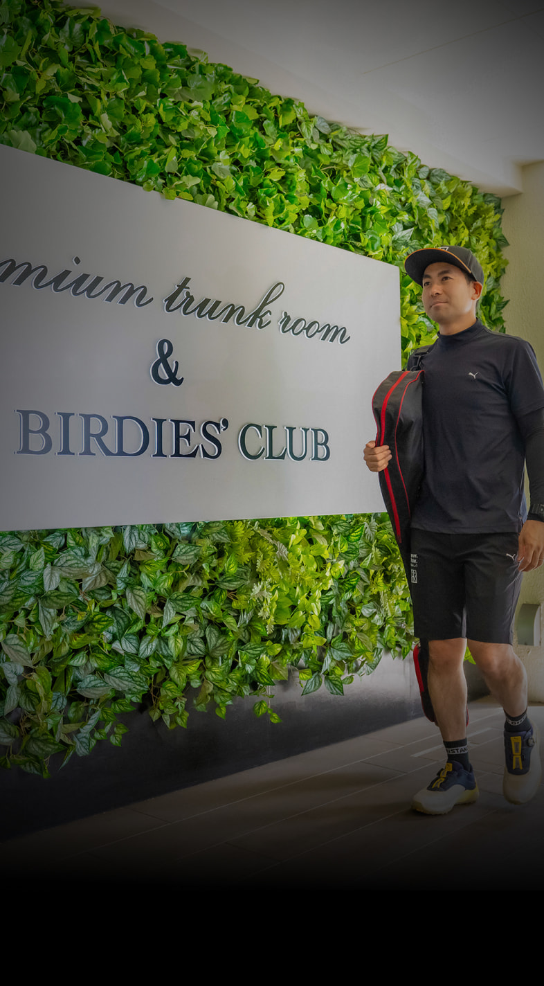 A person in athletic attire walks past a green wall with a sign reading "Premium Trunk Room & Birdies' Club," holding a red and black bag.