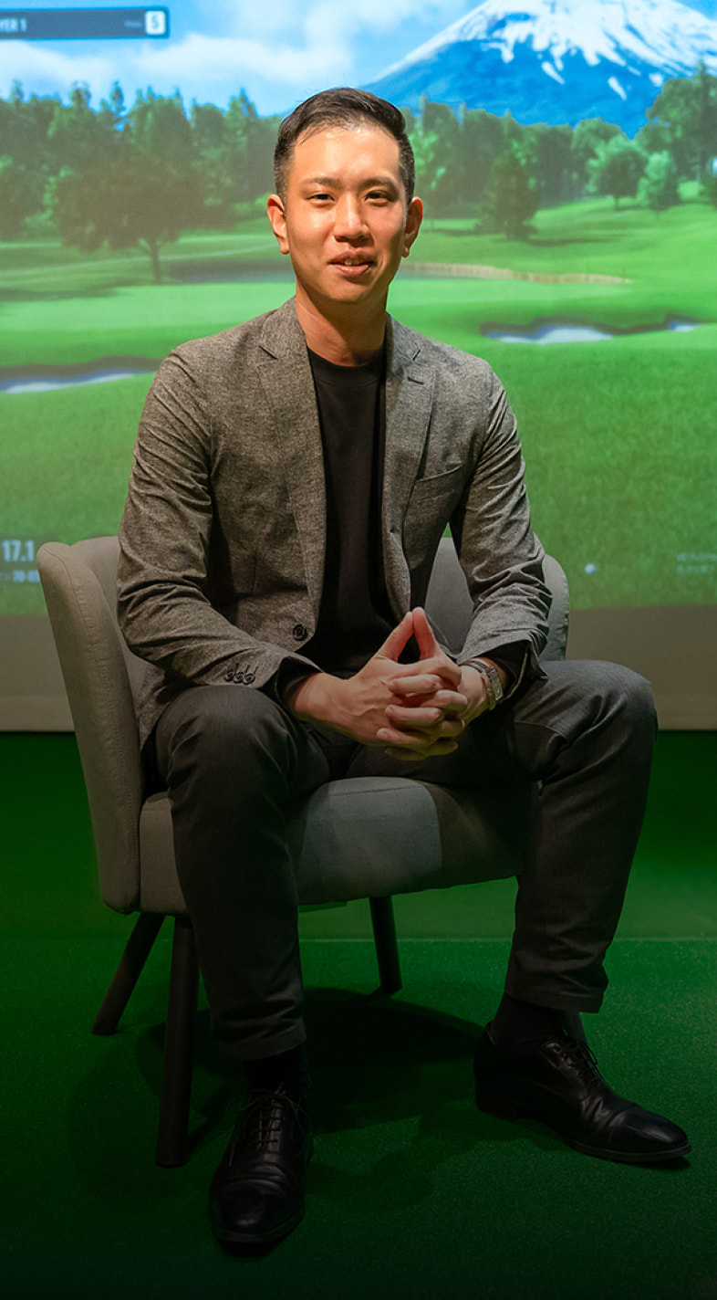 Man sitting on a chair in a golf simulator room, with a virtual golf course displayed on the screen behind him.