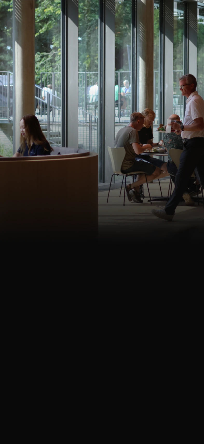People working and dining in a modern space with large windows, featuring green trees outside.