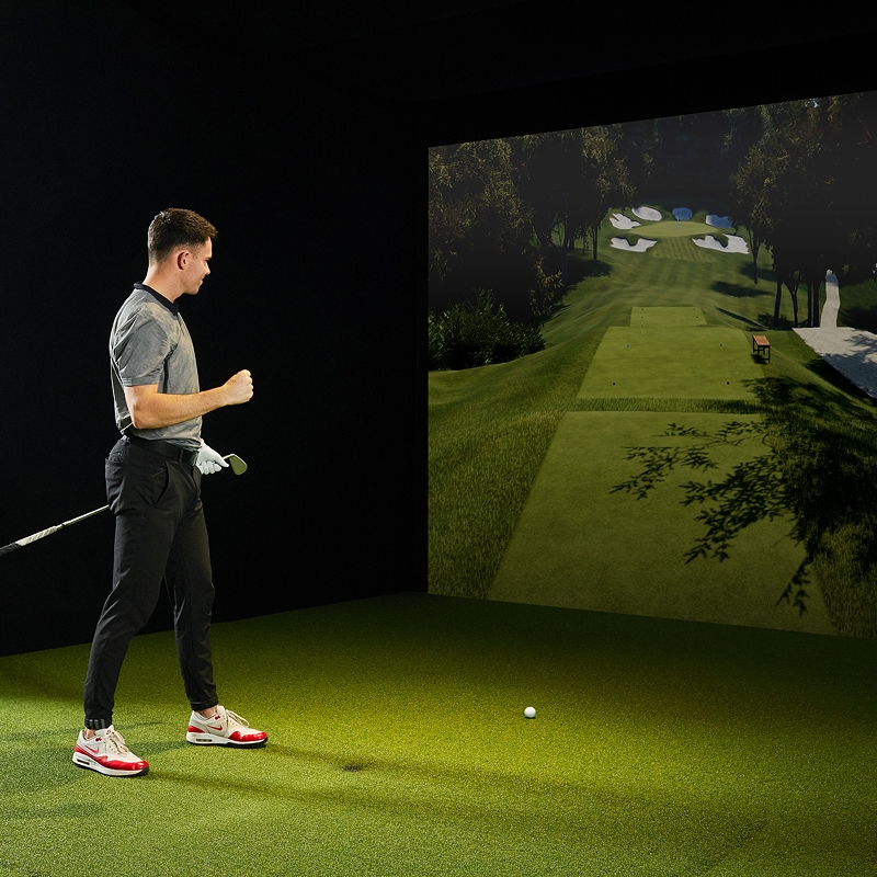 Person playing indoor golf, standing on green turf, facing a large screen displaying a golf course.