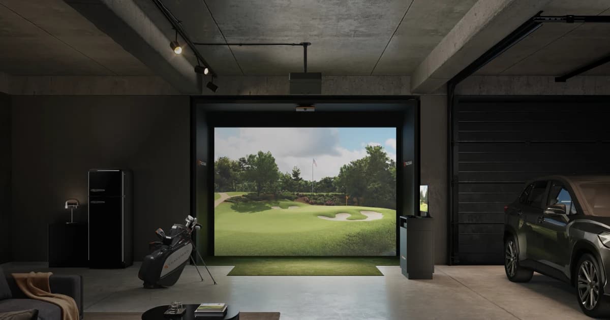 Indoor golf simulator setup in a garage, featuring a large screen displaying a golf course, a golf bag, and a parked car nearby.