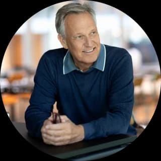 Smiling older man in a blue sweater sitting at a table, holding glasses, with a blurred background of a bright room.