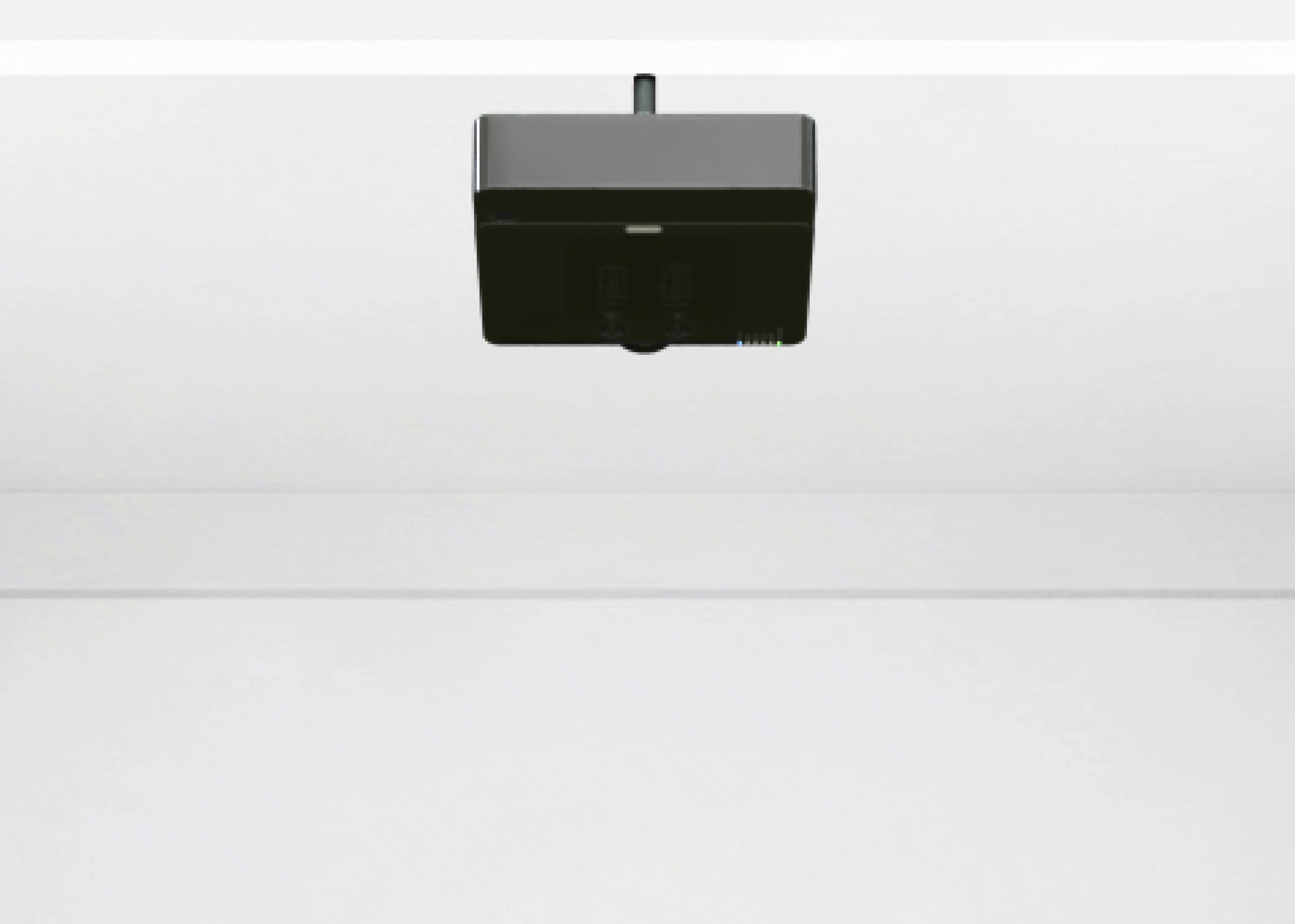 Black ceiling-mounted device with a central lens in a clean, white room.