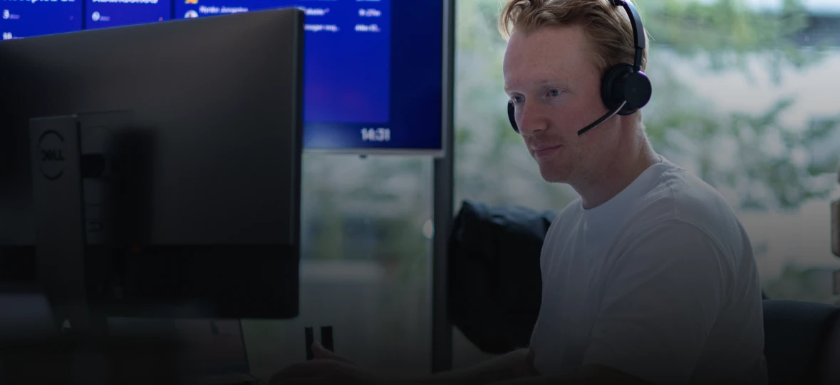 Person with a headset working at a computer, blue screen and greenery visible in the background.