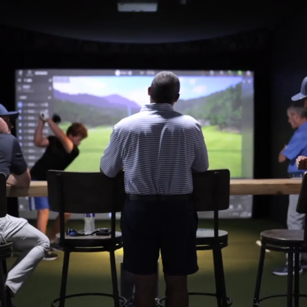 People in an indoor golf simulator; a man in a striped polo stands at a counter watching another player swing at a projected course.