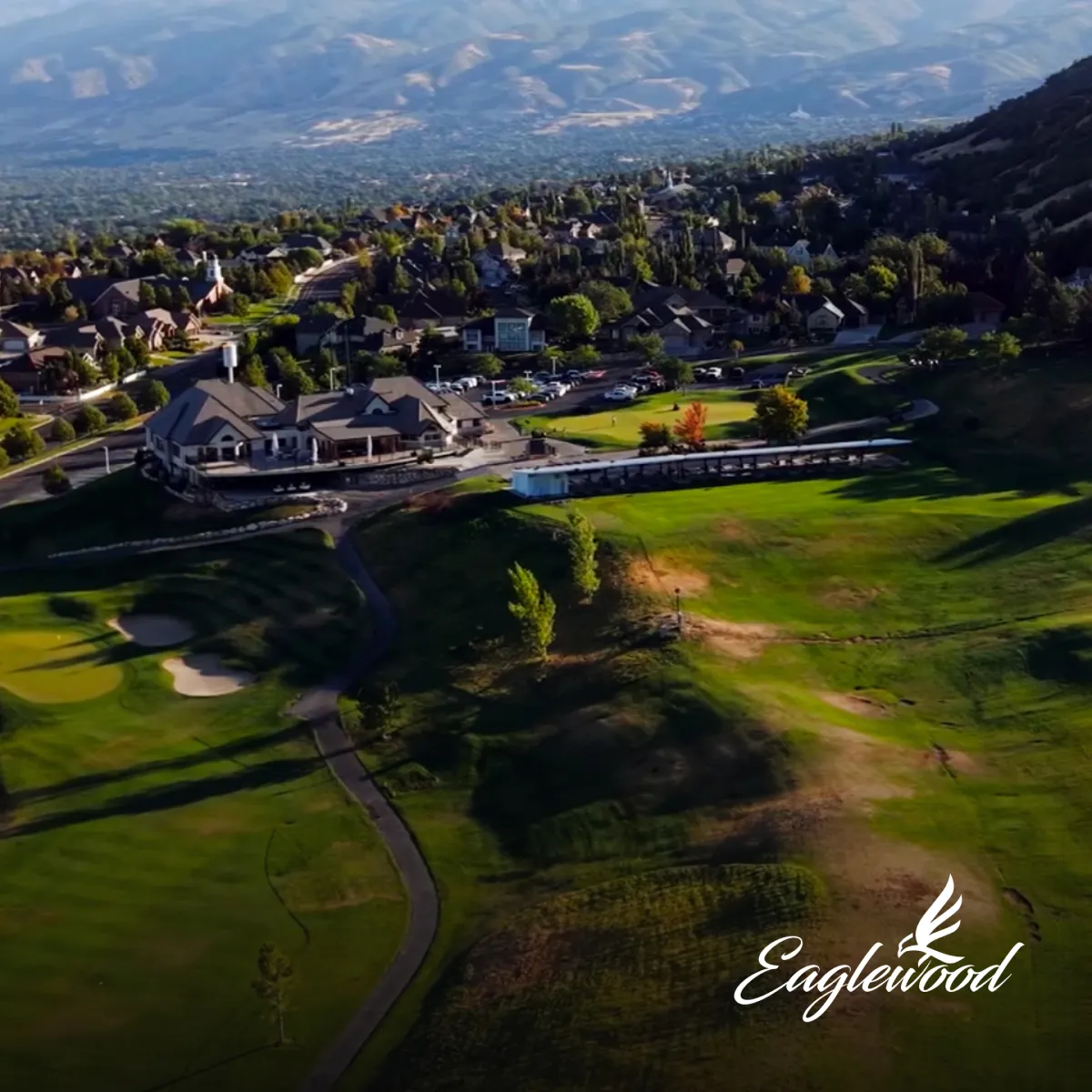 Aerial view of a golf course with a clubhouse, surrounded by houses and hills, under a clear blue sky.