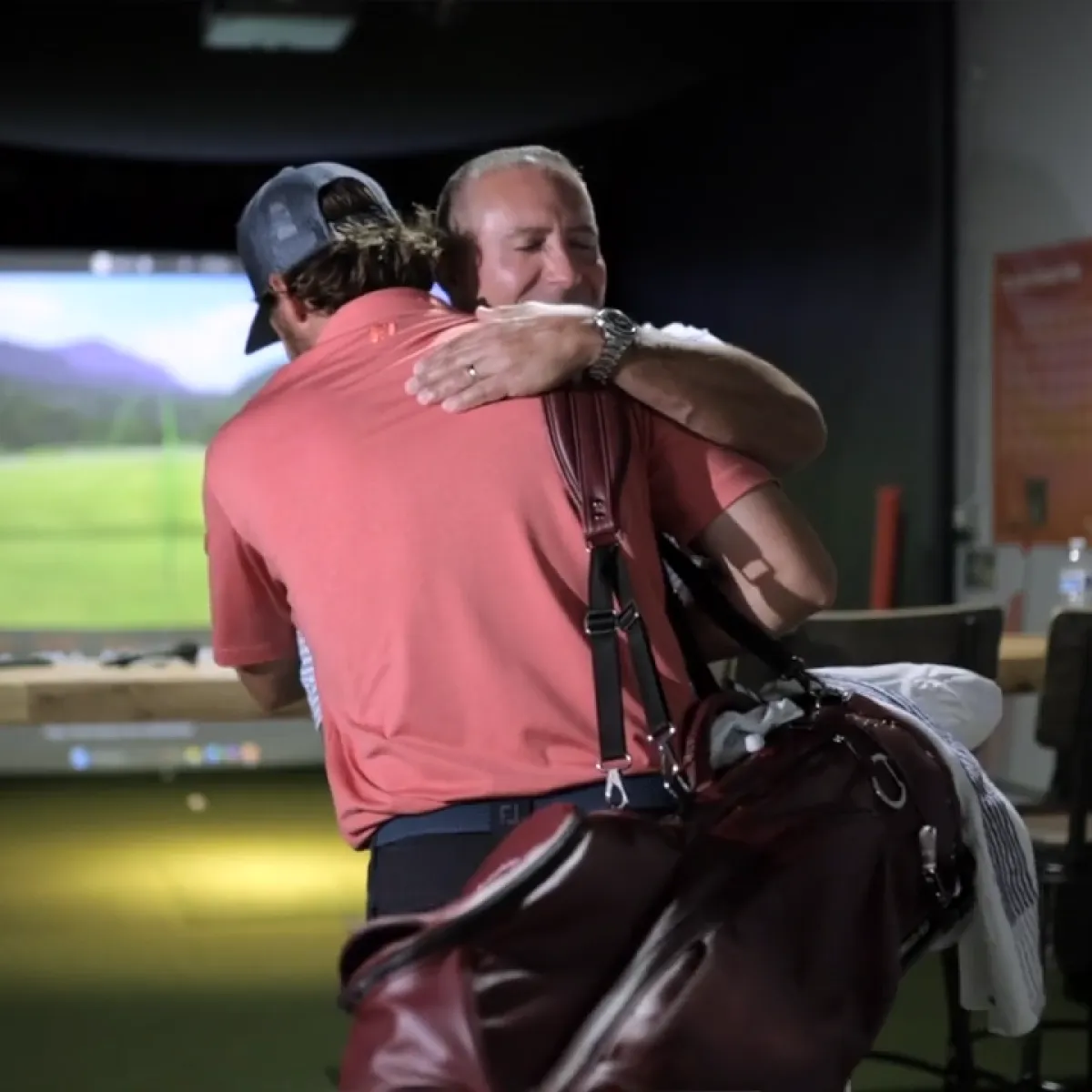 Golfer in a coral shirt and cap, carrying a golf bag, hugs a smiling older man inside an indoor golf simulator.