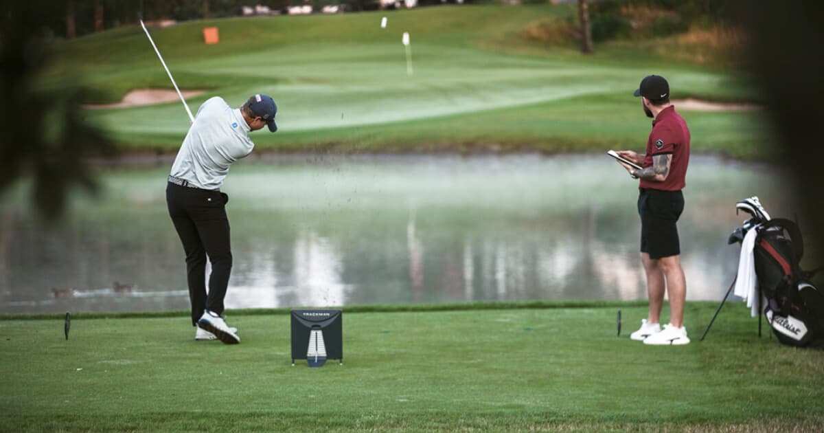 Golfer in mid-swing on a lush course near a pond, with a caddie observing and holding a notepad.