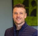 Smiling man with short hair wearing a dark quilted jacket, standing indoors with a green and black abstract artwork in the background.