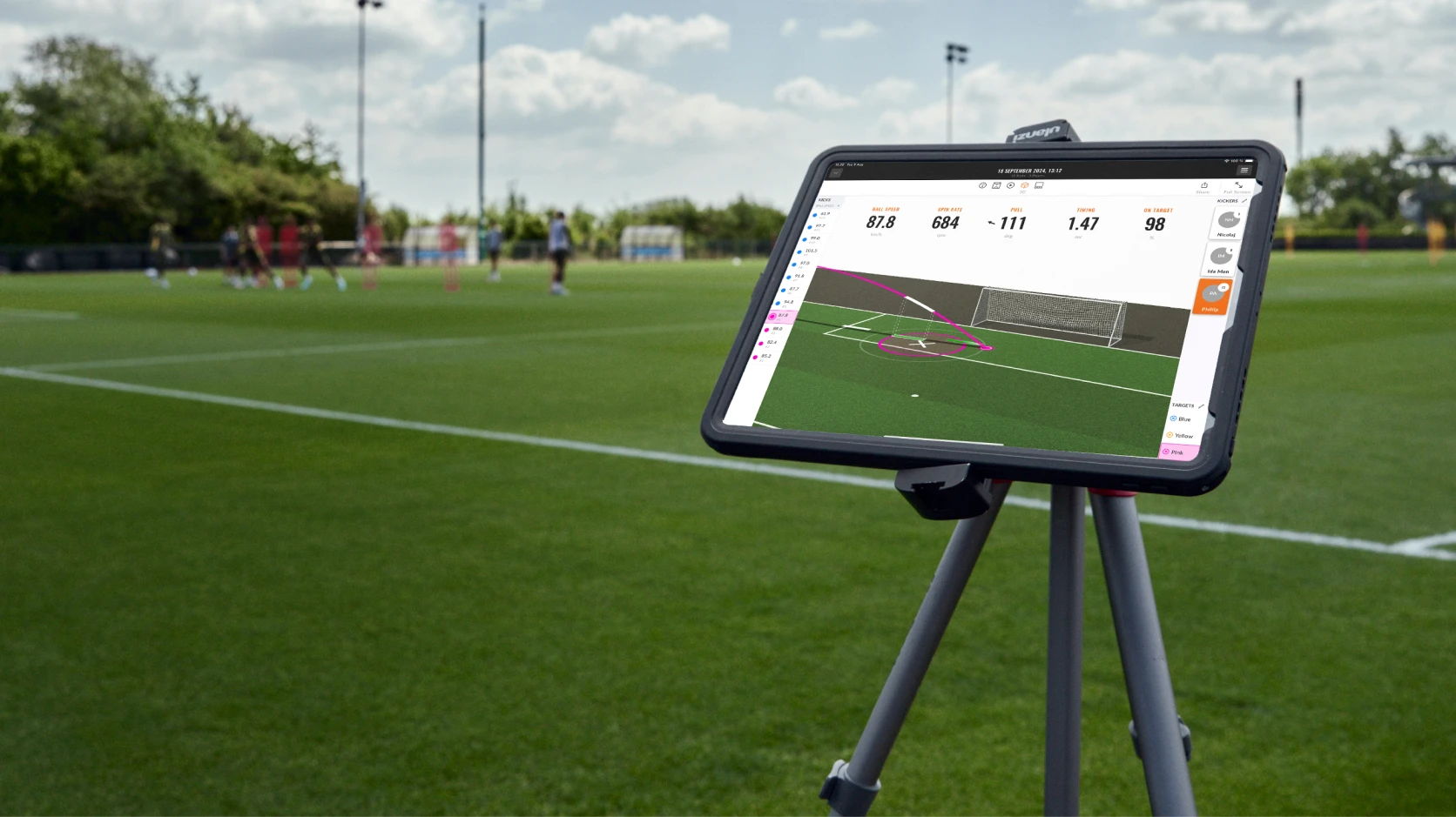 Tablet on tripod displays soccer analytics on field with players training in the background.