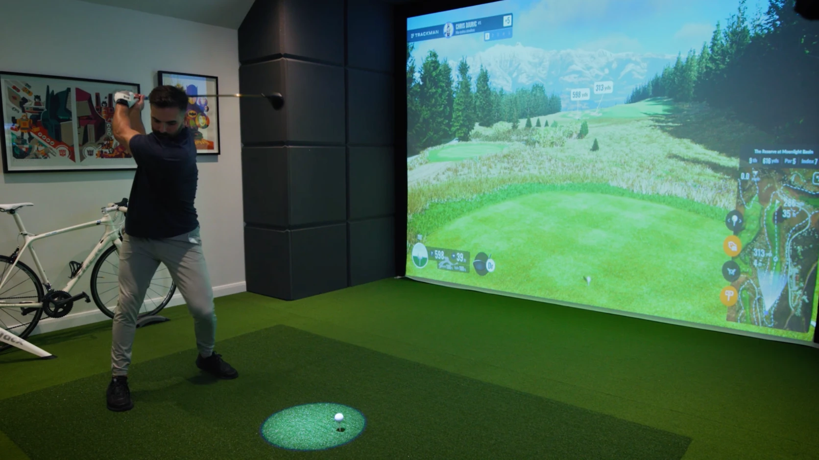 Person swinging a golf club in an indoor simulator room, next to a bicycle, with a virtual golf course projected on the screen.