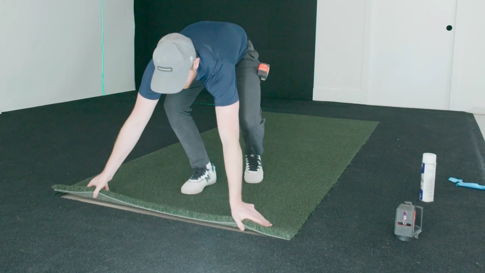 A person in a cap lays down a piece of green turf on a black mat, with tools and adhesive nearby.