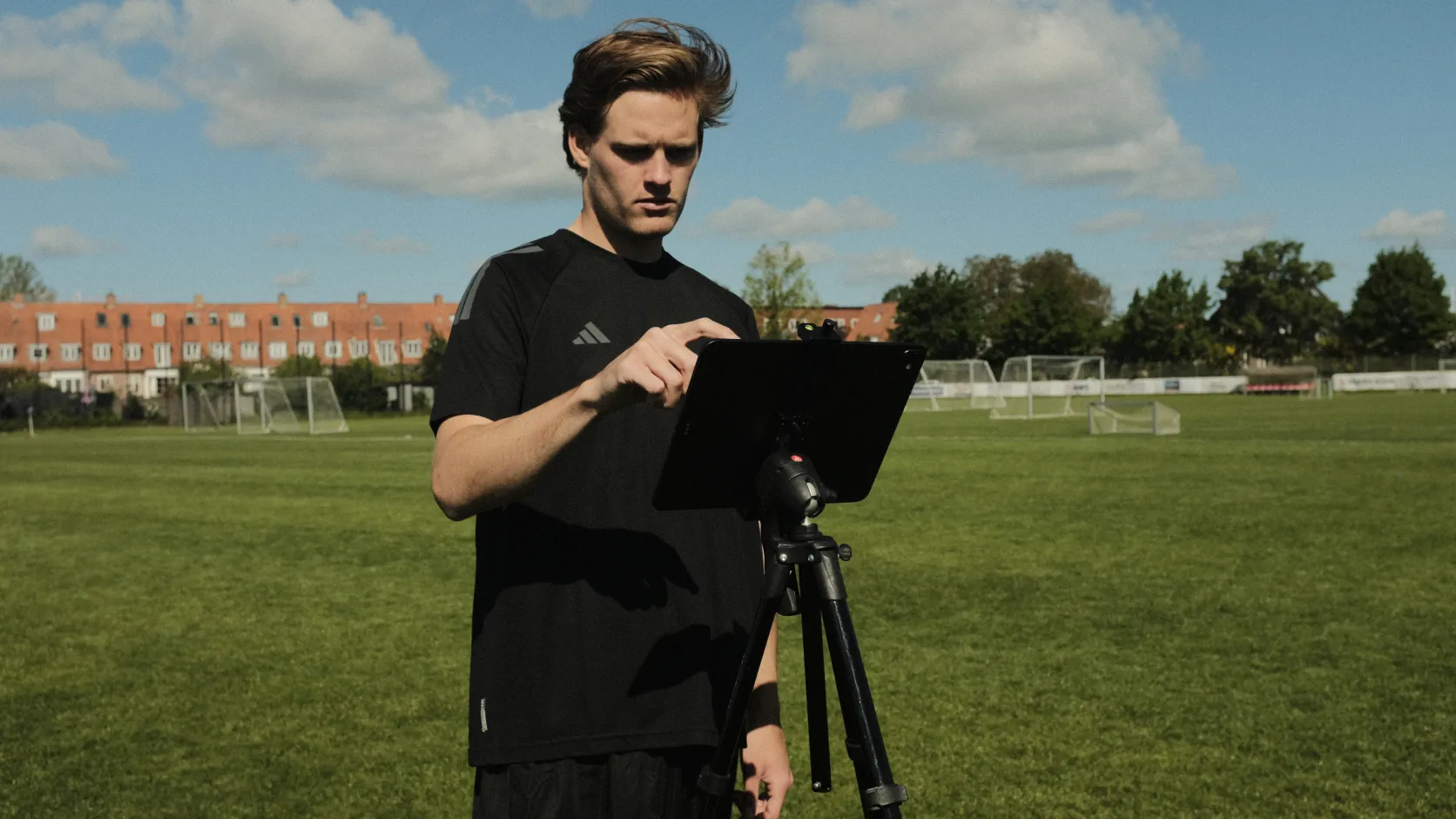 A person in a black sports outfit uses a tablet on a tripod in a sunny soccer field with goals and buildings in the background.