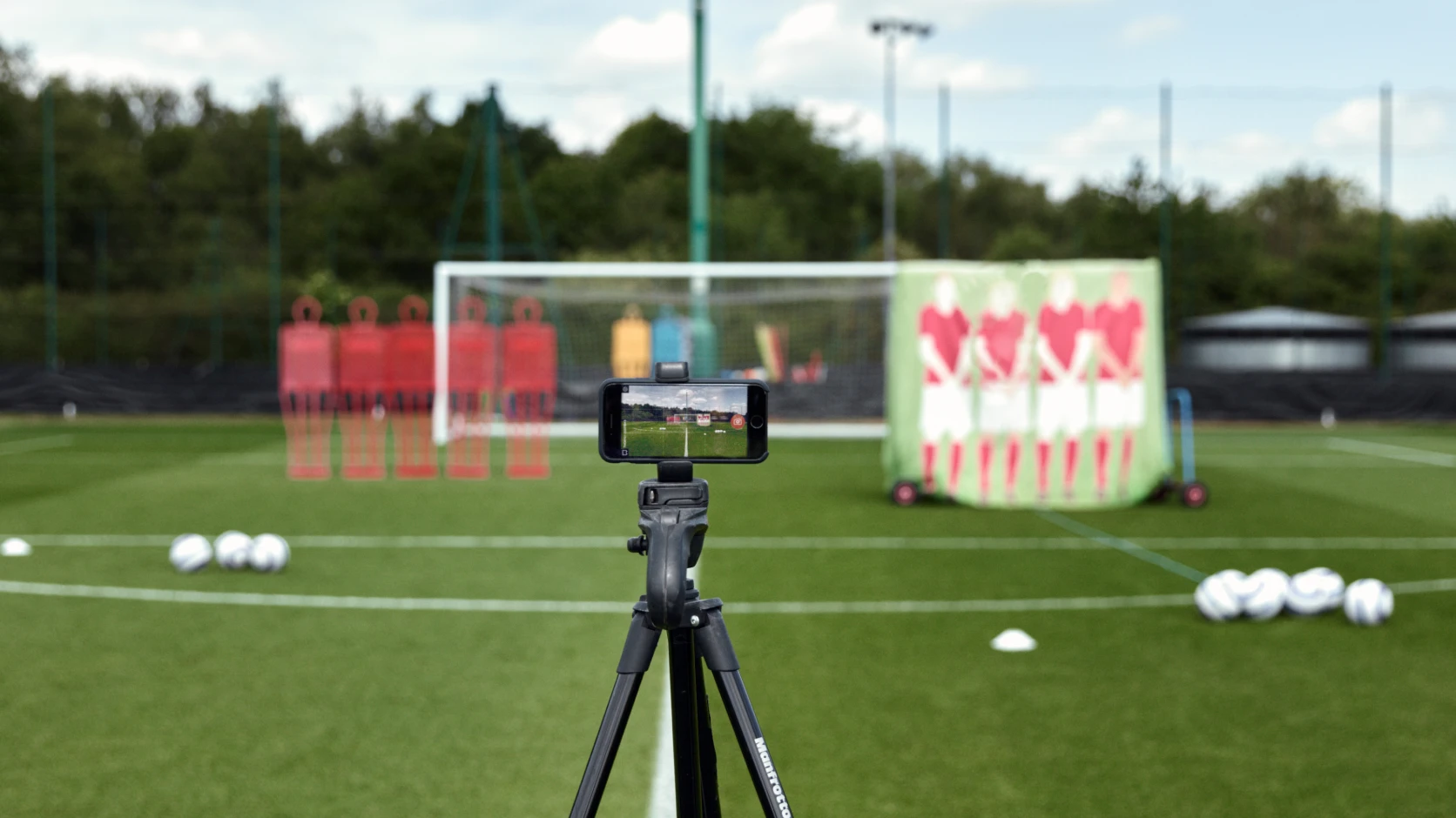 Smartphone on tripod recording a soccer practice setup with training dummies, balls, and cones on a grassy field.