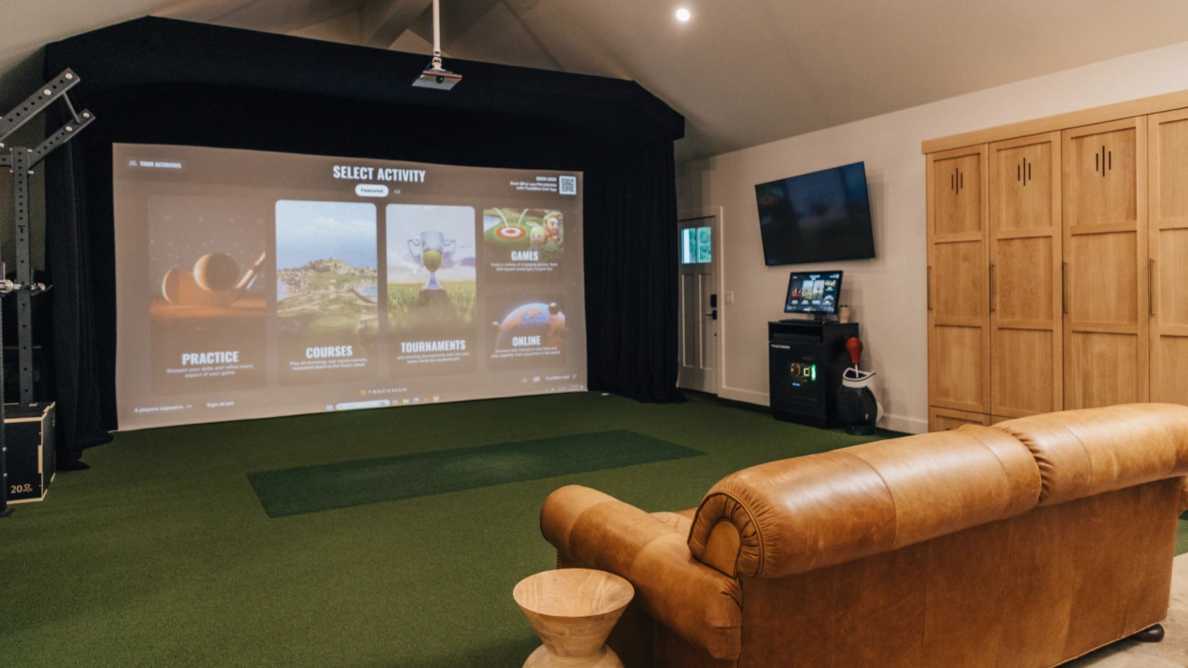 Home golf simulator room with a large projector screen, leather couch, wooden cabinets, and golf equipment on green carpet.