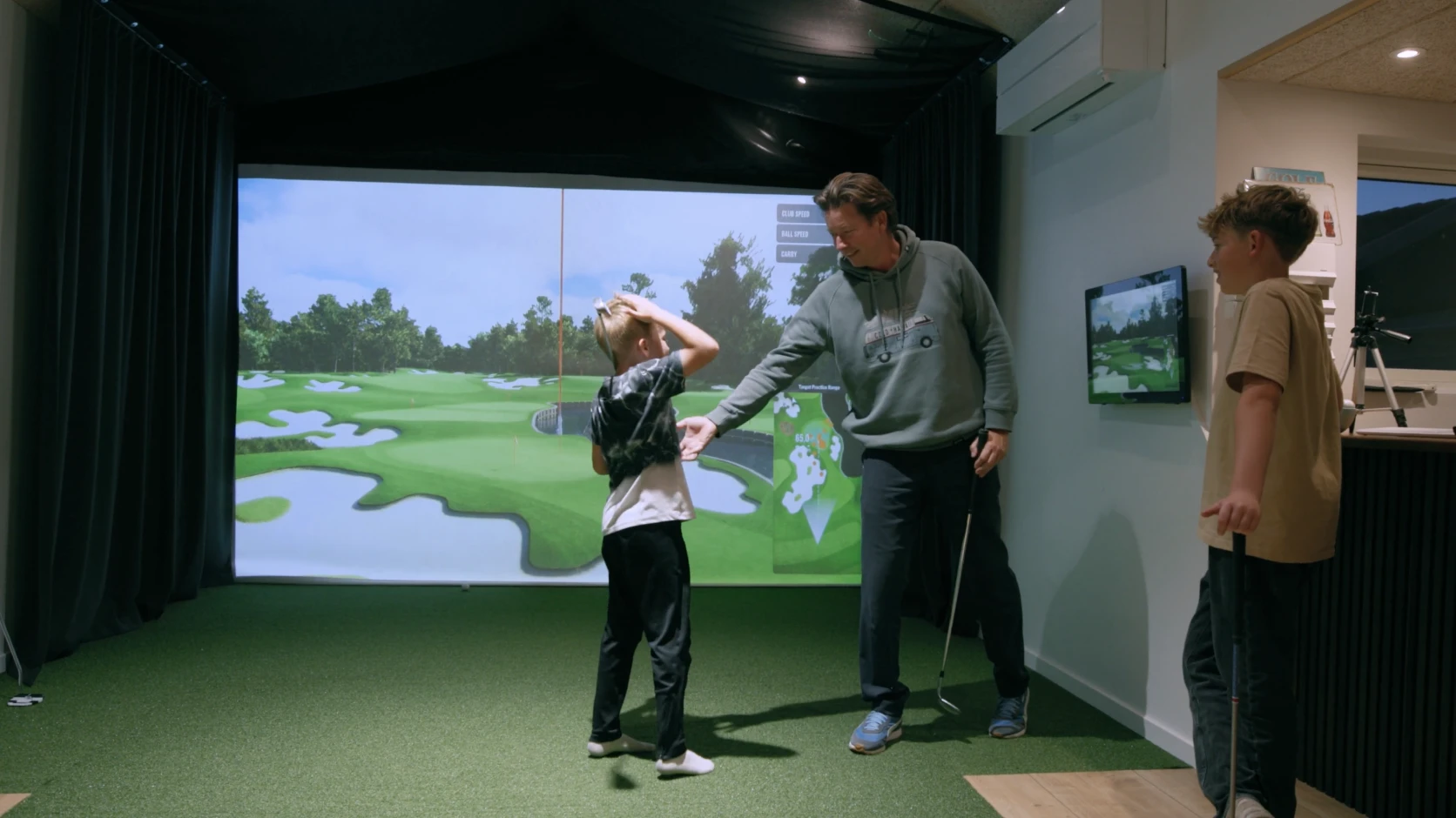 Man and two kids in a golf simulator room; one boy holds his head, the other stands with a club. Screen displays a golf course image.