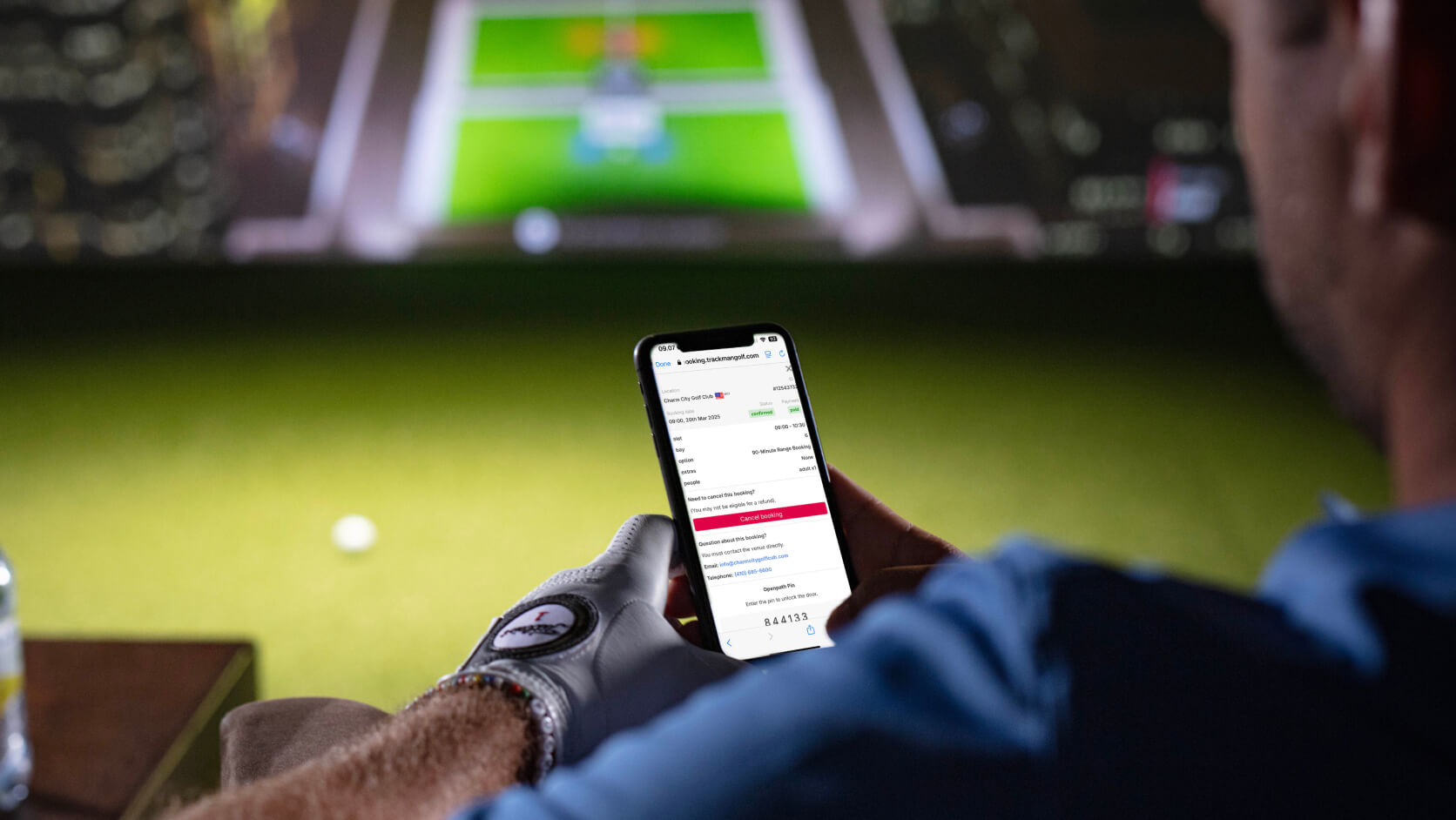 Person in a golf glove using a smartphone with a Trackman Golf and golf simulator in the background