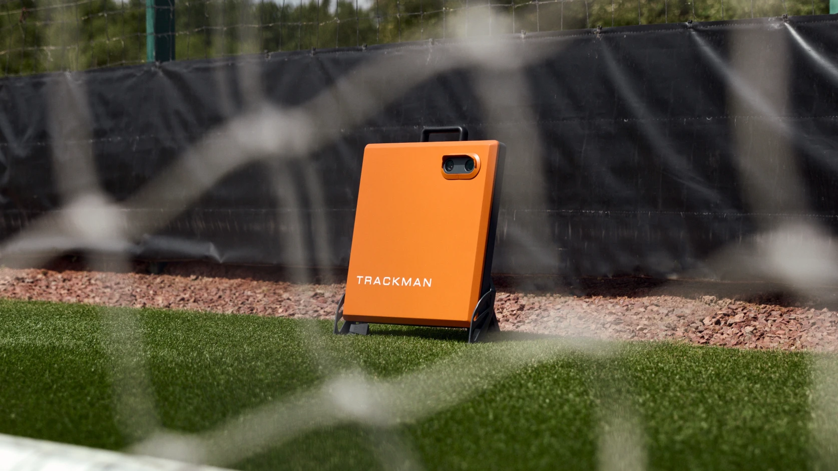 Orange TrackMan device on a sports field near a fence, seen through a net.