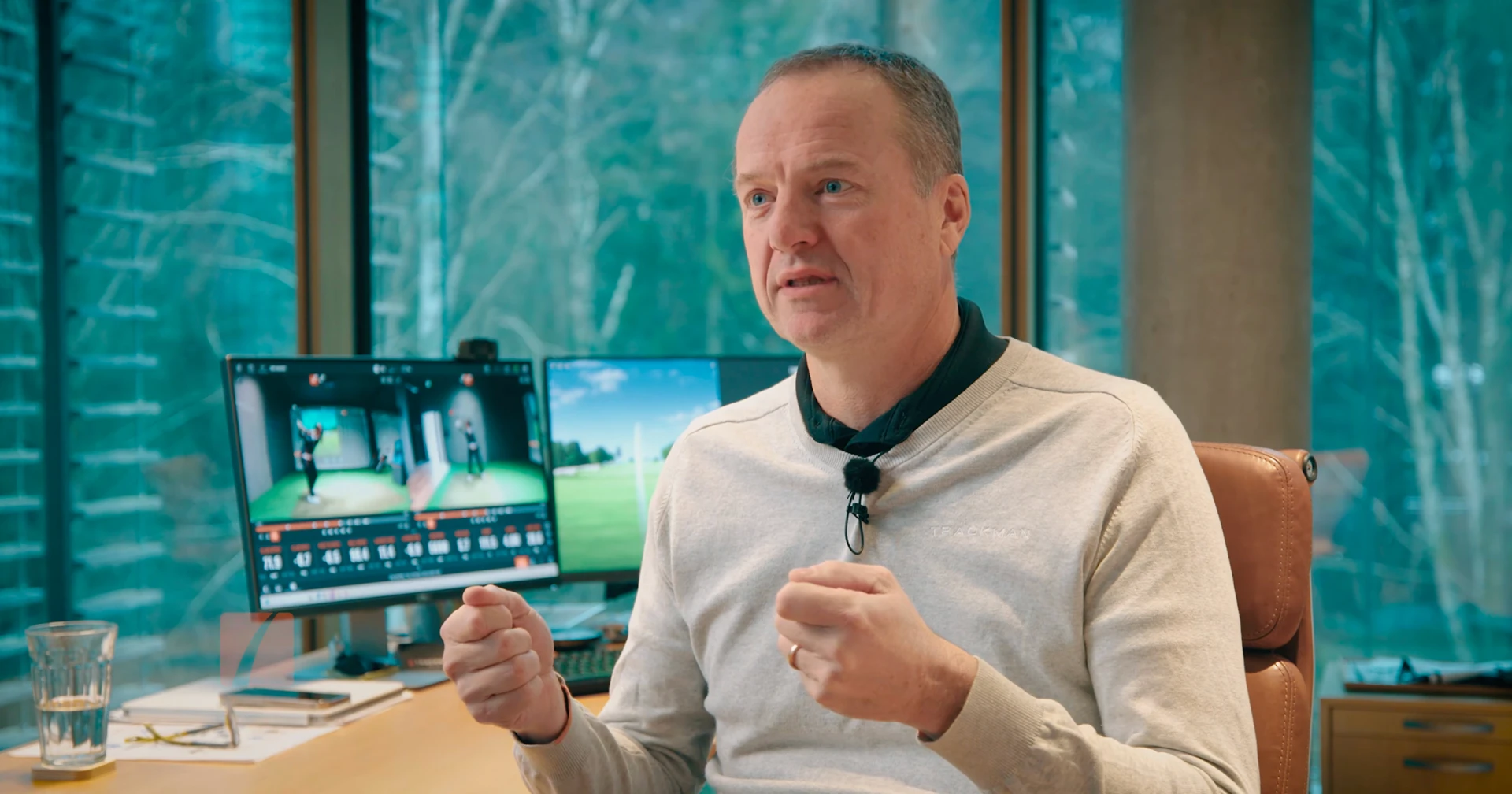 Man sitting in an office, gesturing while talking, with computer screens behind him displaying sports footage and data.