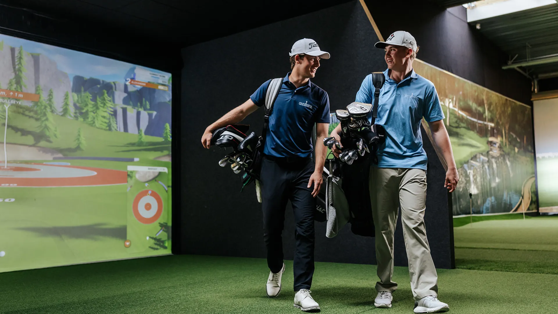 Two golfers with bags walk indoors on artificial turf, smiling and conversing, with a golf simulator screen displaying a scenic course in the background.