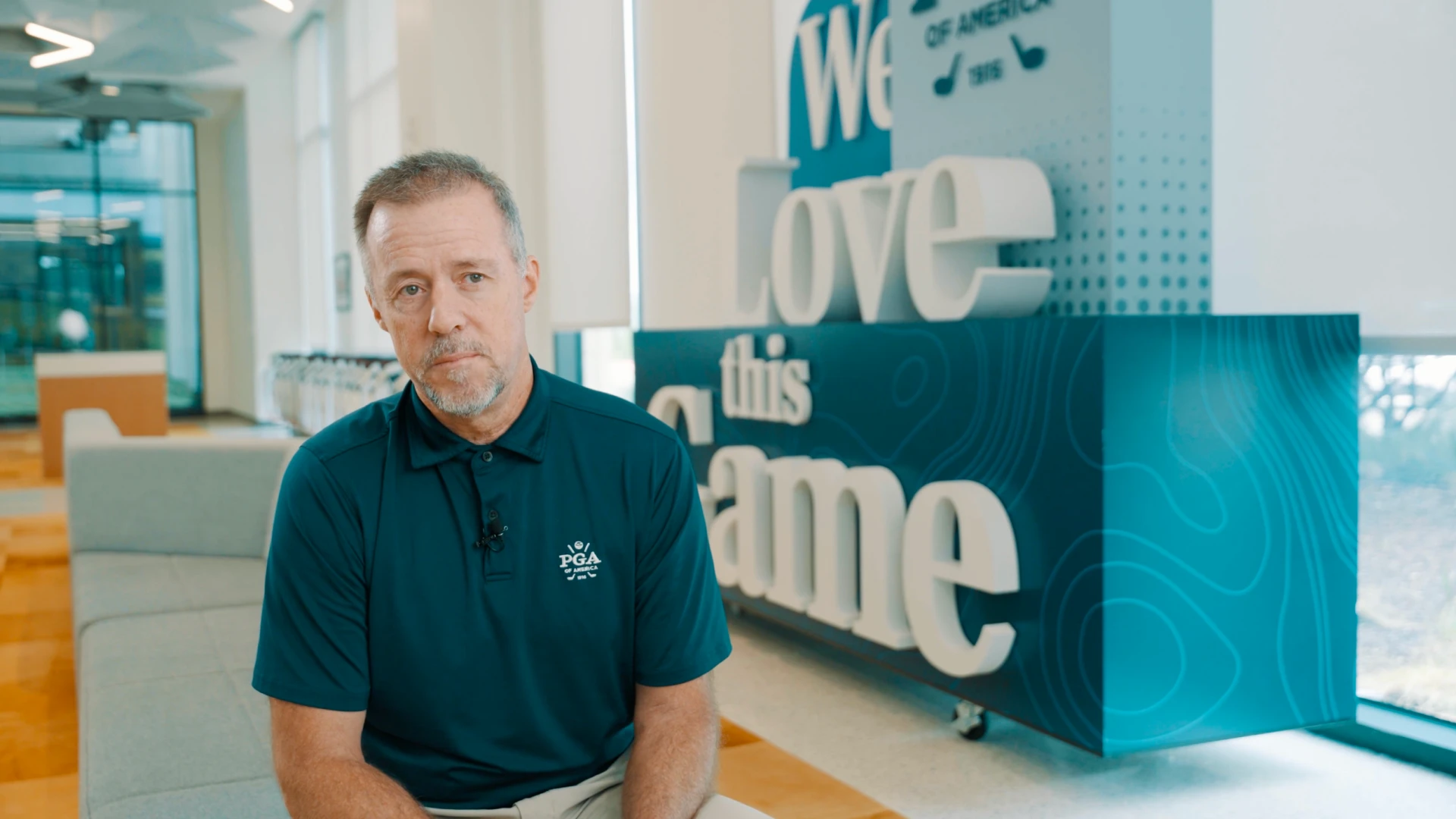 A man in a dark polo shirt sits indoors with a "We Love this Game" sign in the background.