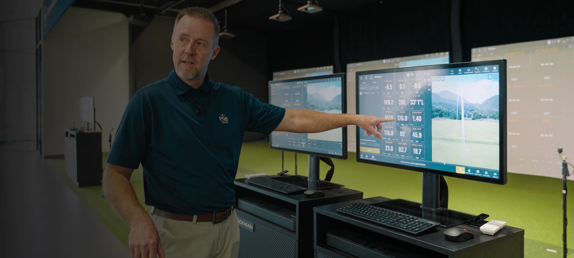 A person in a blue shirt points at data on a large screen with golf simulation metrics. Two monitors and computer equipment are visible.