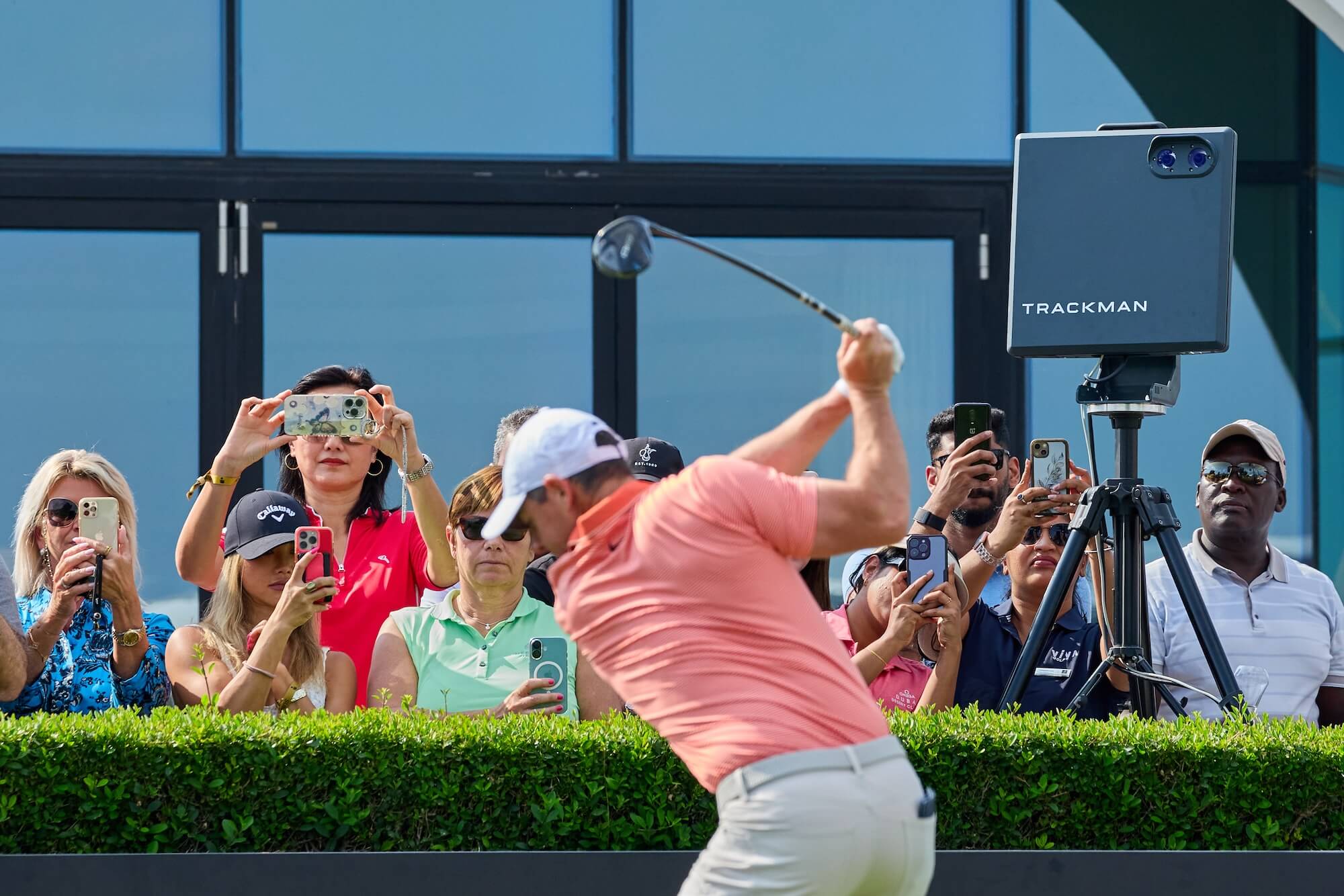 Rory McIlroy swings a club as spectators behind him take photos, with a TrackMan device visible nearby.