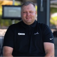 A man in a black polo shirt with a name tag sits outdoors, smiling. The background is blurred, showing a screen and greenery.
