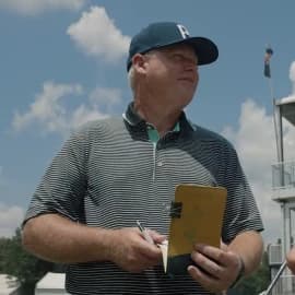 Man in a striped polo and cap holding a notebook and pen, standing outdoors under a blue sky with clouds.