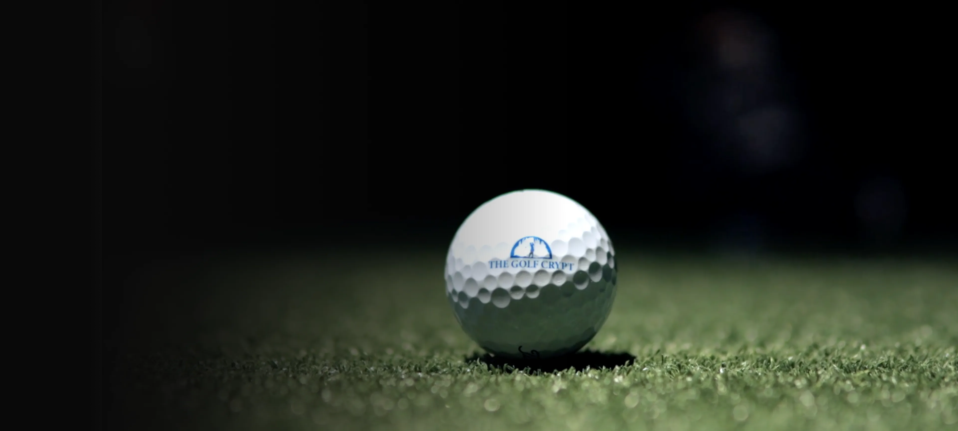 Close-up of a white golf ball with "The Golf Crypt" logo resting on green turf against a dark background.