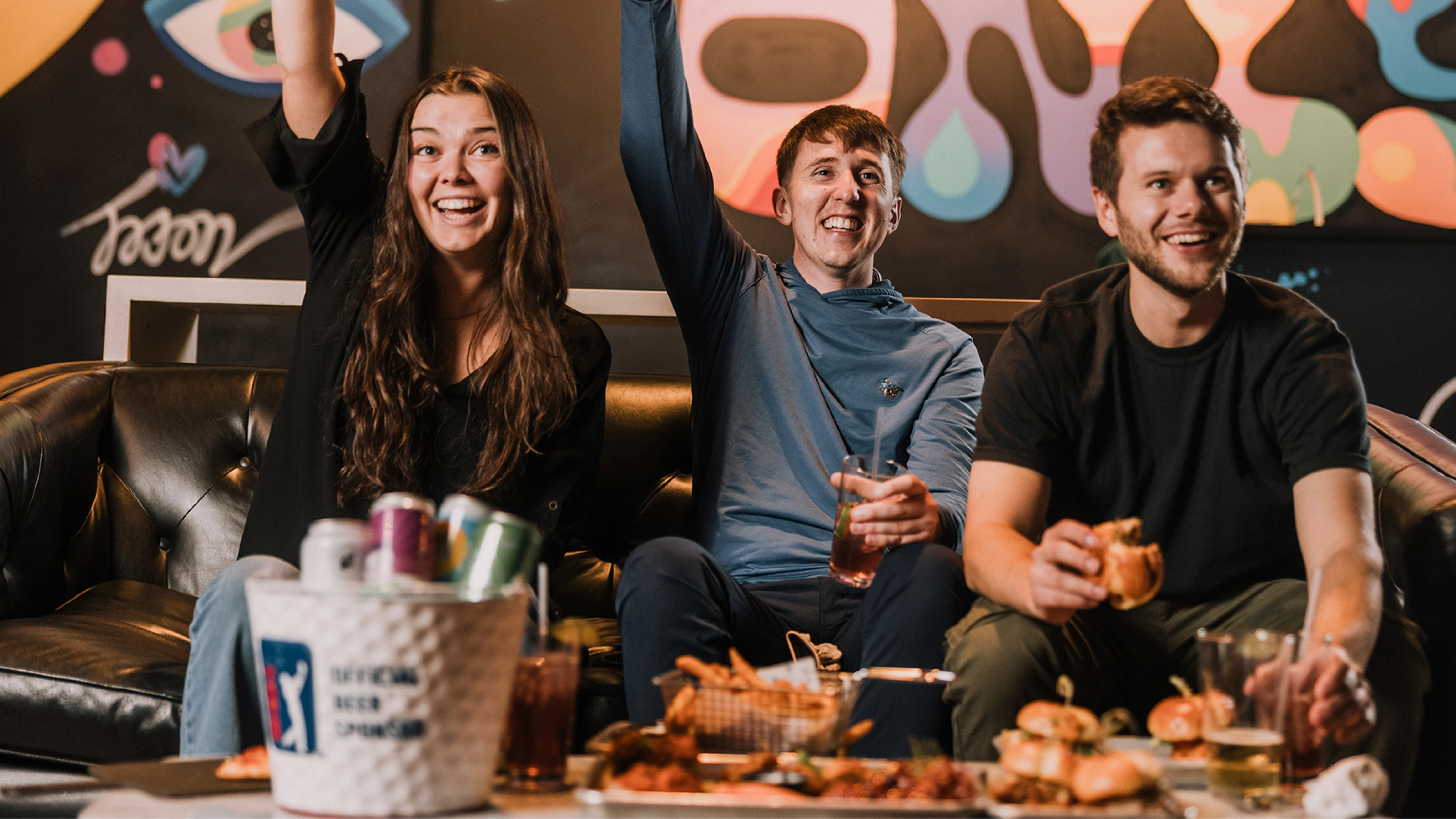 Three people on a sofa cheer enthusiastically while enjoying drinks and snacks. A colorful wall art is in the background.
