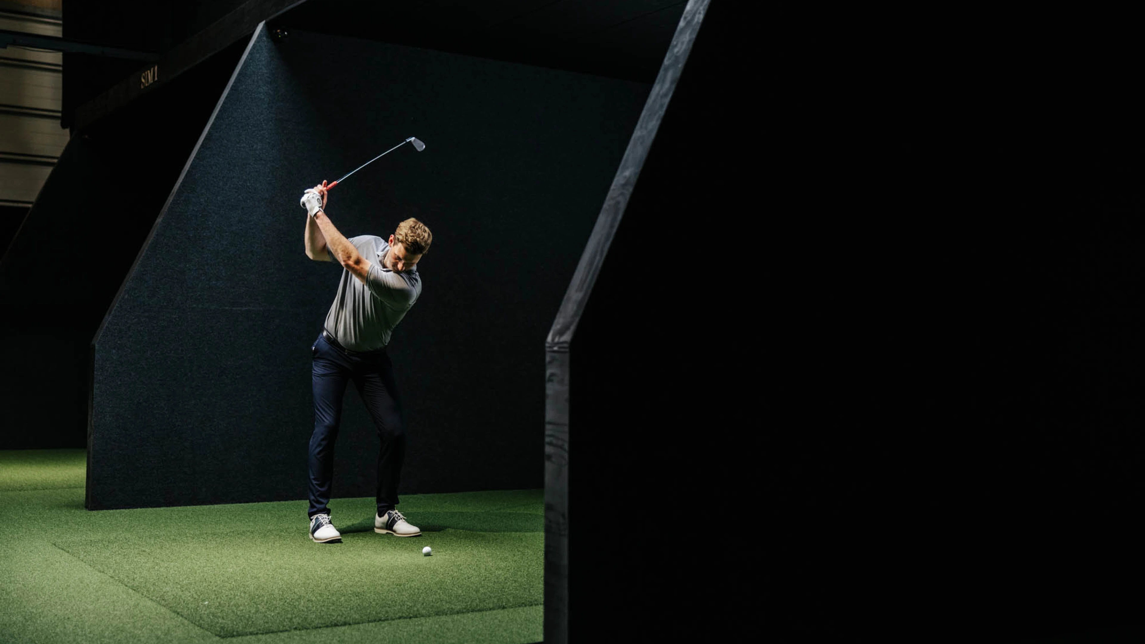A golfer in mid-swing indoors on artificial turf in a dimly lit room, focusing intensely on striking a golf ball.